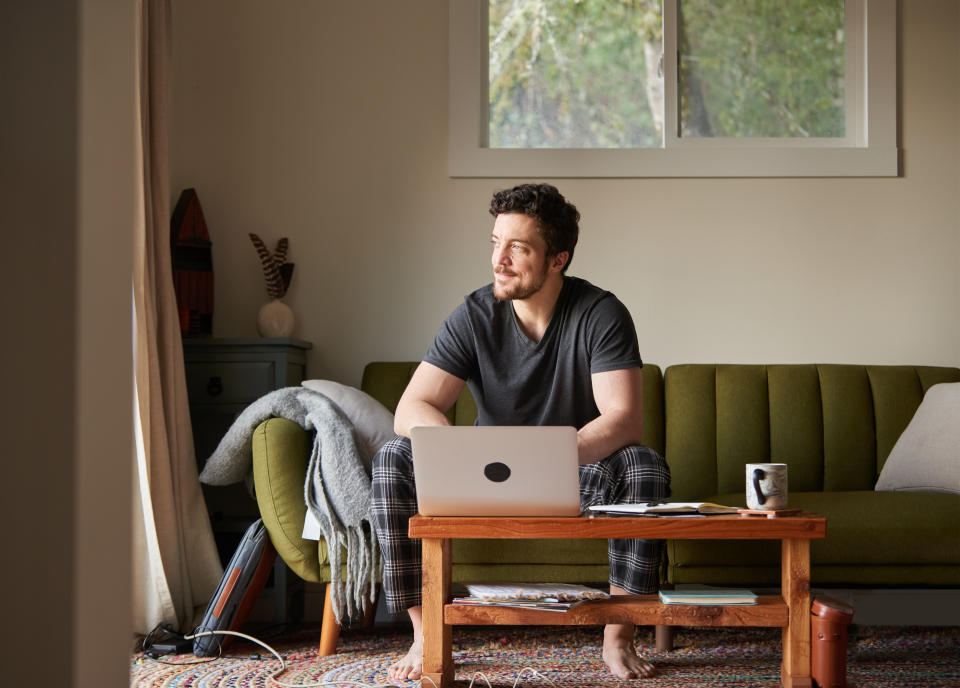 Smiling young man wearing pajamas thinking while working on a laptop on his living room sofa in the morning