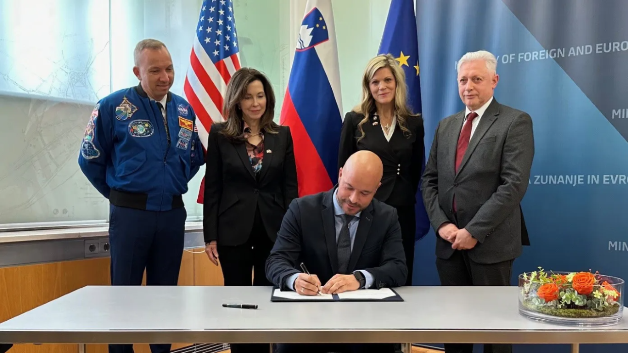  A group of people stand behind a man signing a document in front of flags. 