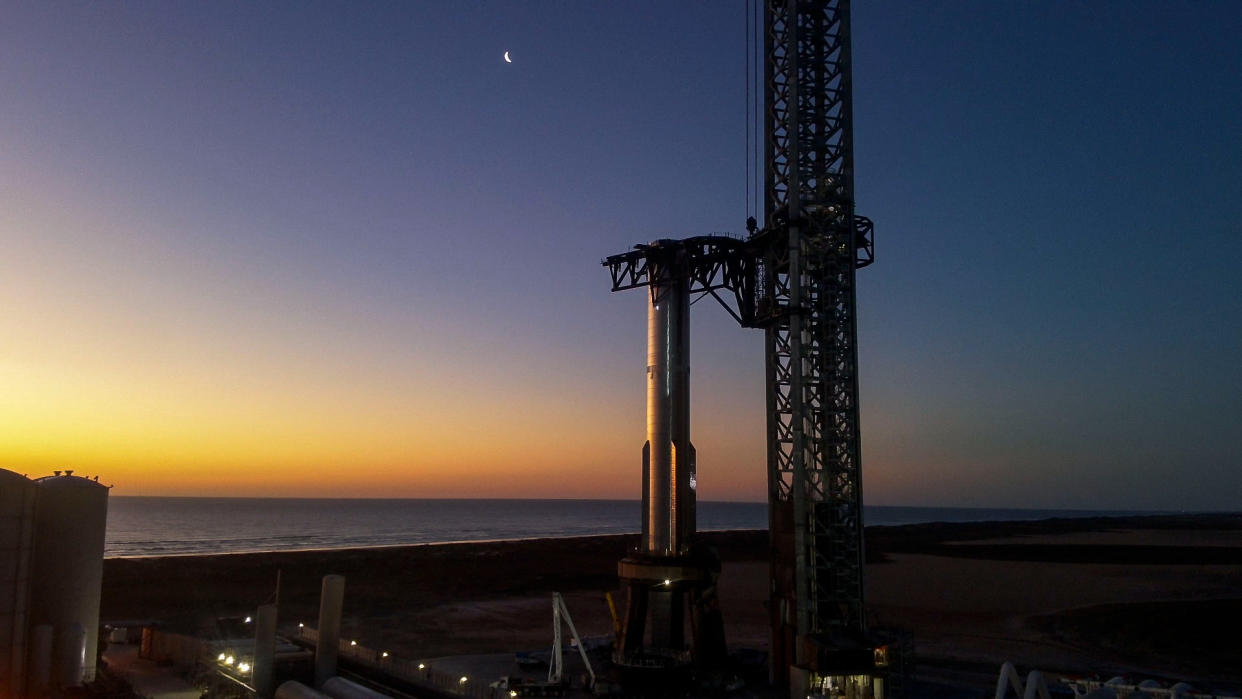  A big silver rocket stands on a launch pad at dawn or dusk, with the ocean in the background. 