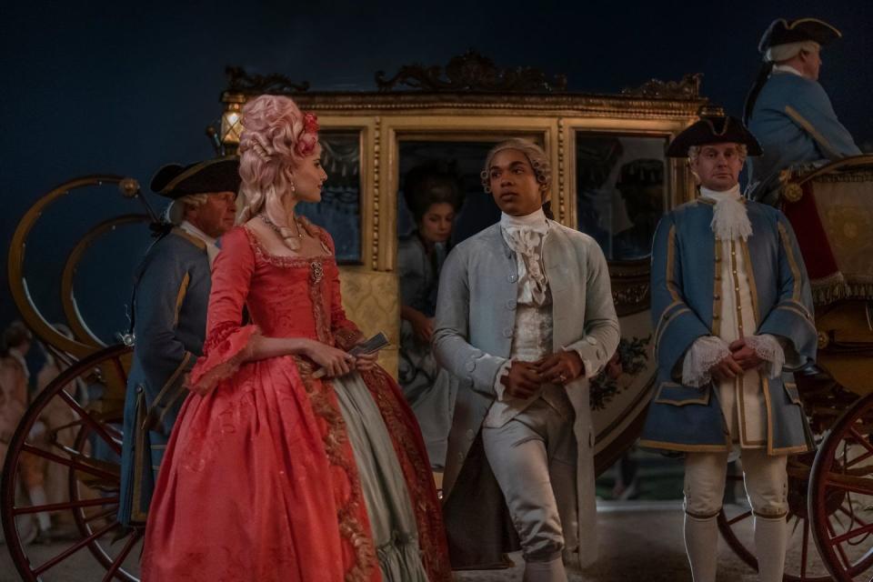 a promotional photo from the film chevalier, featuring actors lucy boynton and kelvin harrison jr in 18th century period costume attire, standing in front of a golden carriage attended to by several people in blue uniforms