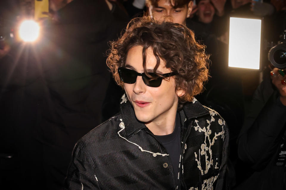 A closeup of Timothée Chalamet wearing sunglasses as fans and paparazzi take his picture