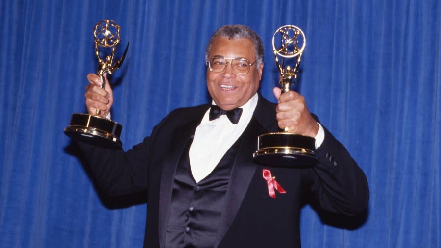 James Earl Jones holds up an Emmy trophy in each hand. He is wearing a black tuxedo with a red ribbon pinned to the lapel.