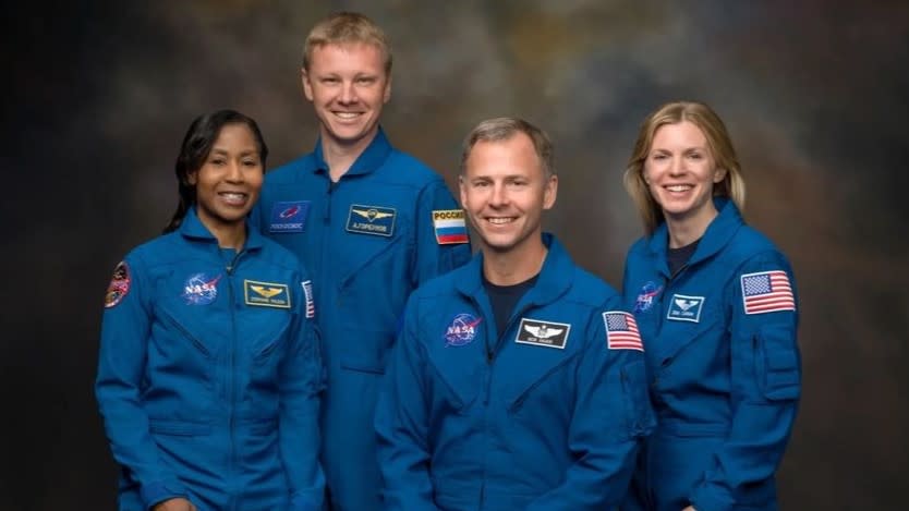  Four smiling people in blue flight suits stand in front of a dark gray-brown background. 