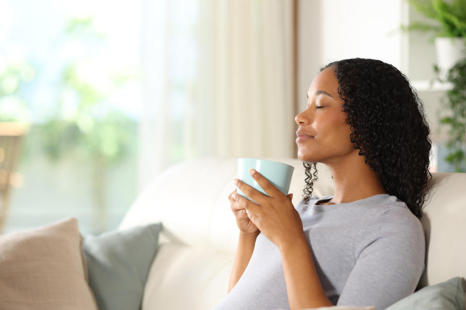 Woman with curly hair sits on a couch, eyes closed, enjoying a drink from a mug with a relaxed expression