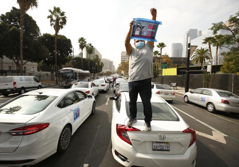 LOS ANGELES, CA - OCTOBER 08: Rideshare driver Jesus Ibarra stands on his car in support as app based gig workers held a driving demonstration with 60-70 vehicles blocking Spring Street in front of Los Angeles City Hall urging voters to vote no on Proposition 22, a November ballot measure that would classify app-based drivers as independent contractors and not employees or agents, providing them with an exemption from California's AB 5. The action is part of a call for stronger workers' rights organized by the Mobile Workers Alliance with 19,000 drivers in Southern California and over 40,000 in all of California. Los Angeles on Thursday, Oct. 8, 2020 in Los Angeles, CA. (Al Seib / Los Angeles Times