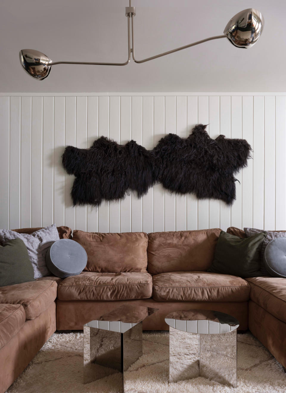 A living room with white panelled walls, brown sofa, mirror side tables and polished nickel ceiling light