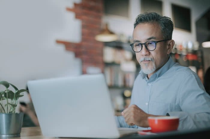 An older man uses his laptop to read the news while sitting in a coffee shop.