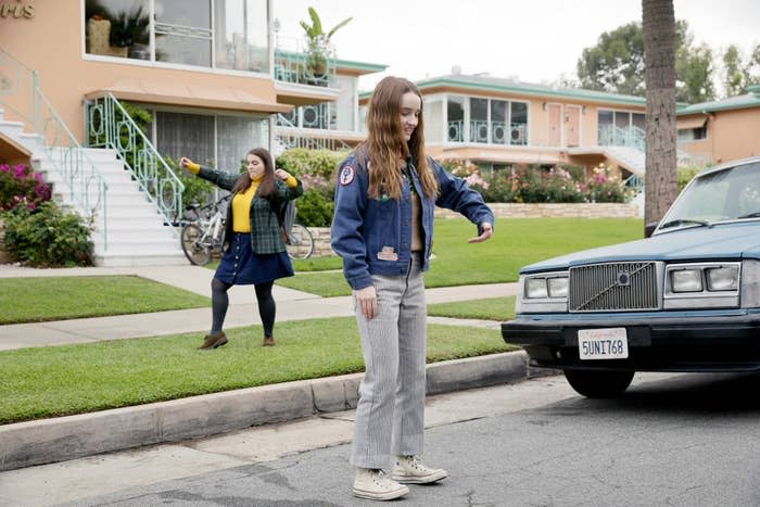 Kaitlyn Dever and Beanie Feldstein are pictured in a casual neighborhood scene from the movie "Booksmart." Beanie dances in the background while Kaitlyn stands near a car