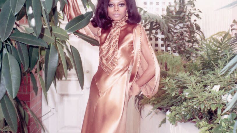 diana ross stands among plants while wearing a silky golden floor length dress