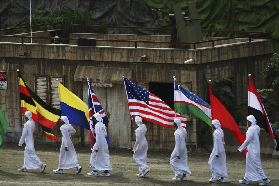 Emirati boys holding flags of the participating countries at an arms show march past a theatrical machine gun position at the opening ceremony of the International Defense Exhibition and Conference in Abu Dhabi, United Arab Emirates, Sunday, Feb. 17, 2019. The biennial arms show in Abu Dhabi comes as the United Arab Emirates faces increasing criticism for its role in the yearlong war in Yemen. (AP Photo/Jon Gambrell)