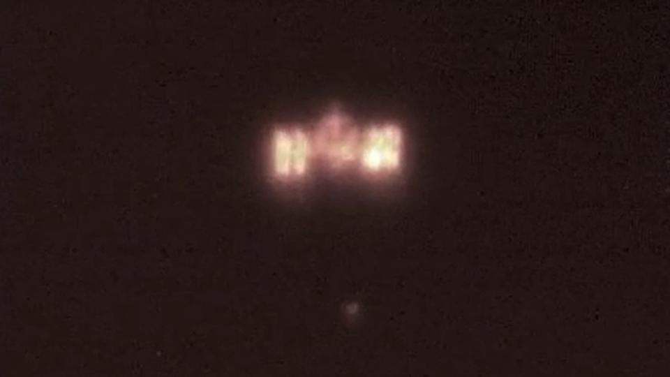  An H-shaped space station glows in reddish orange against a black background as a fuzzy orange dot approaches it. 