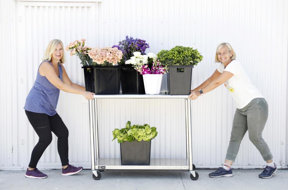 Two women hold a cart with flowers on it.