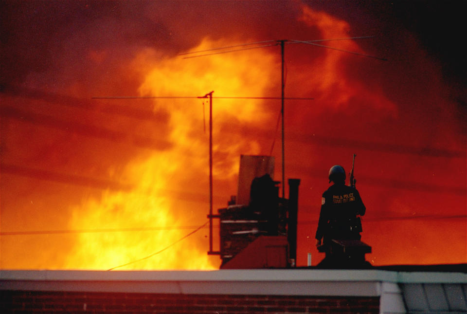 FILE - In this May, 1985 file photo, a Philadelphia policeman is seen on a rooftop as flames rise from a row of burning homes beyond, in Philadelphia. The fire started when police dropped a bomb onto the house of the militant group MOVE, on May 13, 1985 and fire spread throughout the area. A day after Philadelphia's health commissioner was forced to resign over the cremation of partial remains thought to belong to victims of a 1985 bombing of the headquarters of a Black organization, the city now says those victims' remains were never destroyed. City officials told the victims' family Friday, May 14, 2021 that a subordinate had disobeyed Health Commissioner Thomas Farley’s 2017 order to dispose of the remains. (AP Photo/George Widman, File)