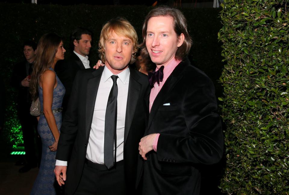 owen wilson and wes anderson, both wearing black suits, smile at a dress party