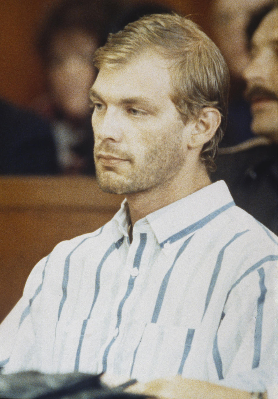 I don't know who is in the image. A person with short hair and a beard wears a striped button-up shirt, seated in what appears to be a formal setting