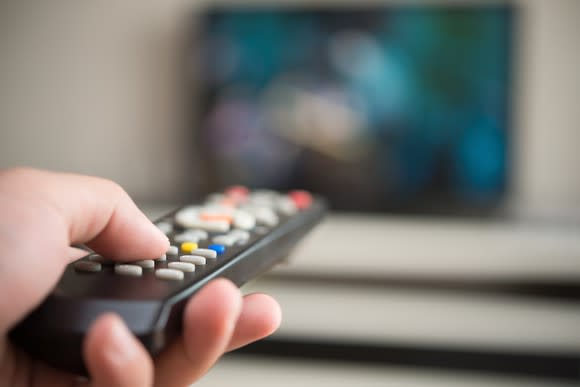 Close-up on a hand holding a TV remote, pointed at a blurry TV set in the background.