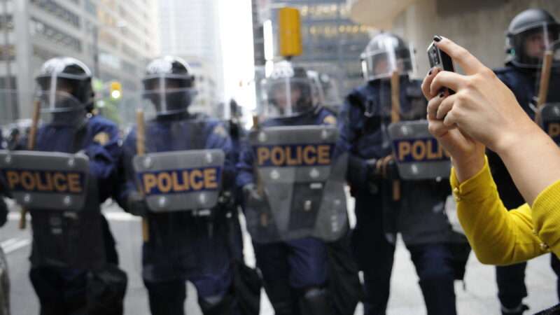 A woman takes pictures on her phone against the backdrop of police in riot gear holding shields.