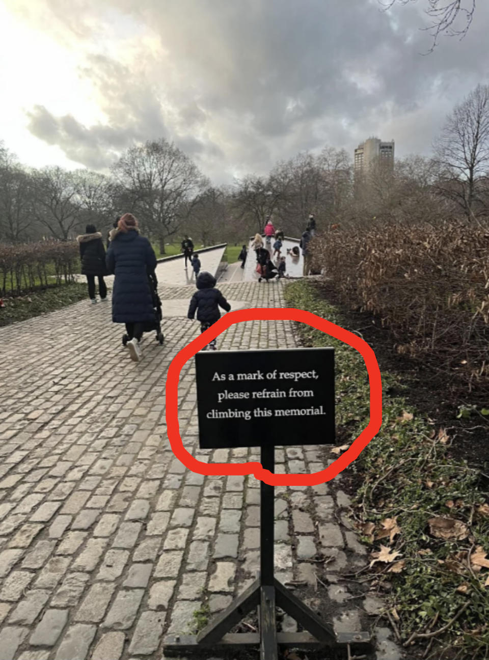 People walk along a cobblestone path with a sign in the foreground reading, "As a mark of respect, please refrain from climbing this memorial."