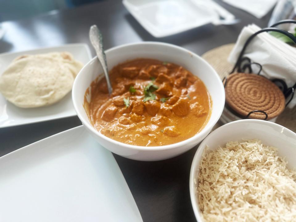 A bowl of curry chicken, with visible chicken pieces and a dark-orange sauce, sits on a table with a white bowl of basmati rice and a white place with naan on it.