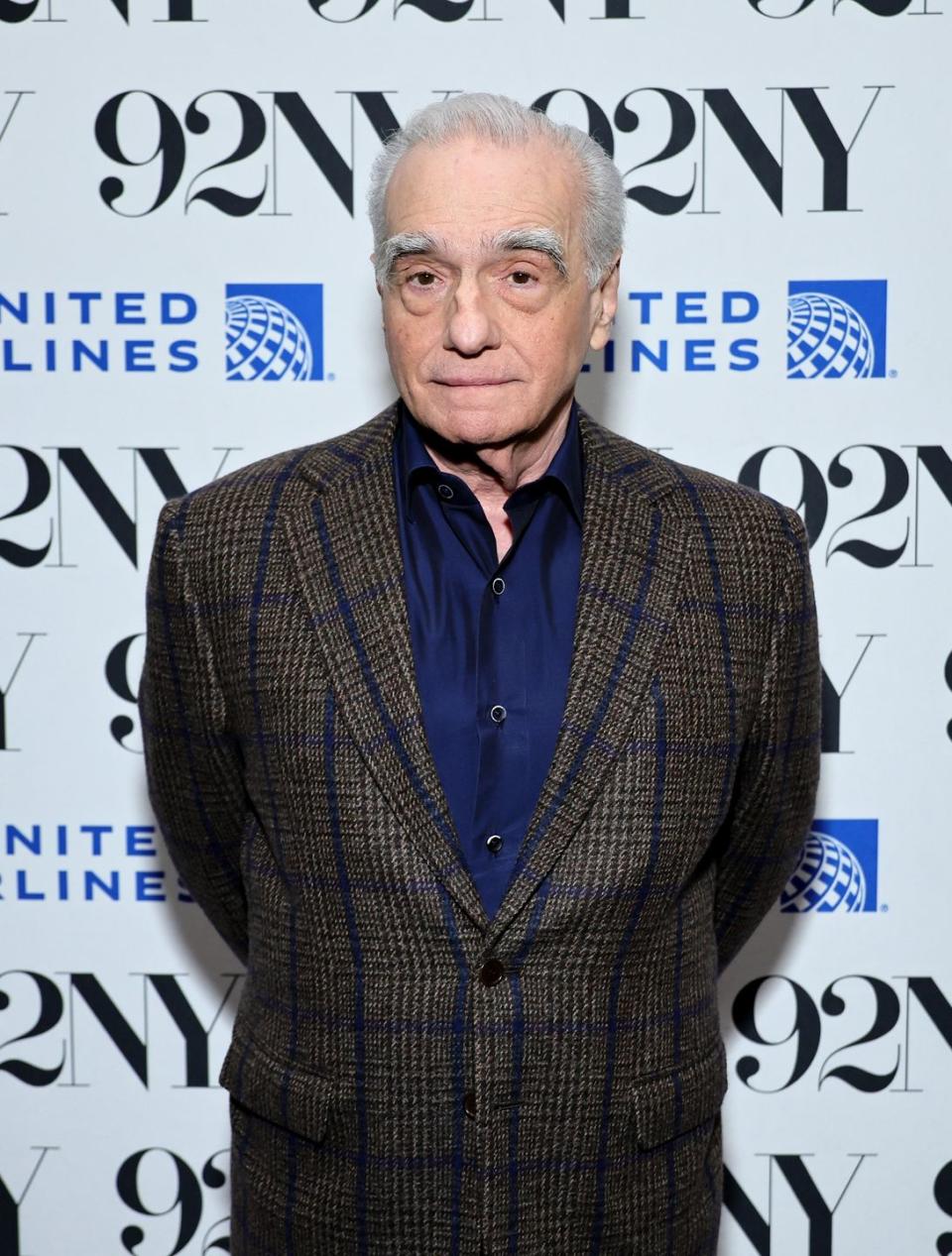 martin scorsese looks at the camera while standing in front of a white background with logos, he weras a brown and blue plaid suit jacket and a dark blue collared shirt