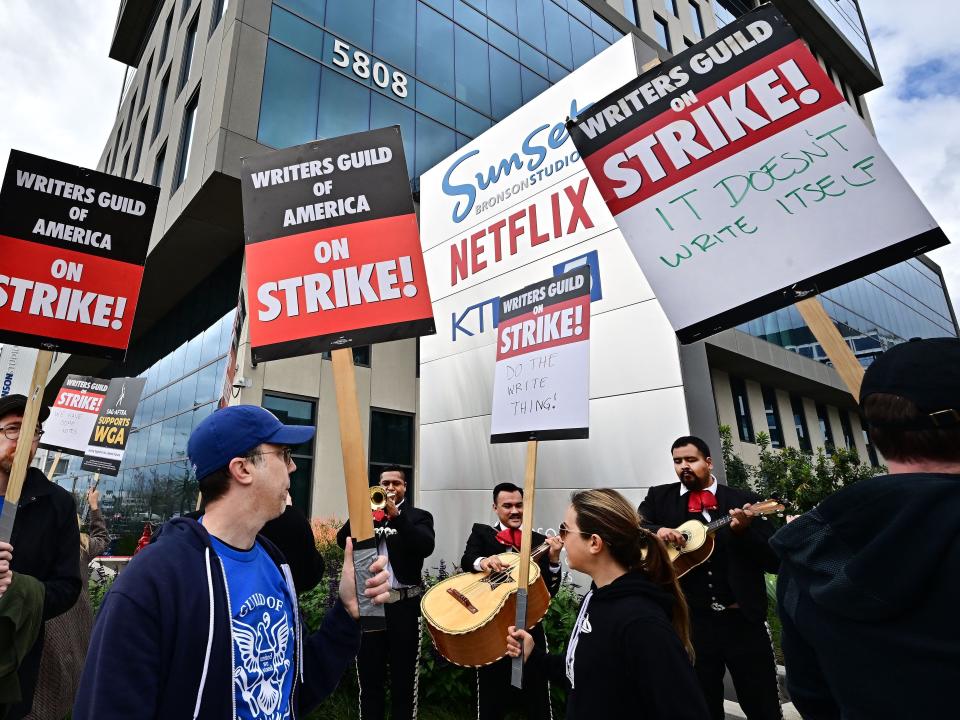 A group of writers on strike hold signs outside of an office building with a Netflix logo in the background.