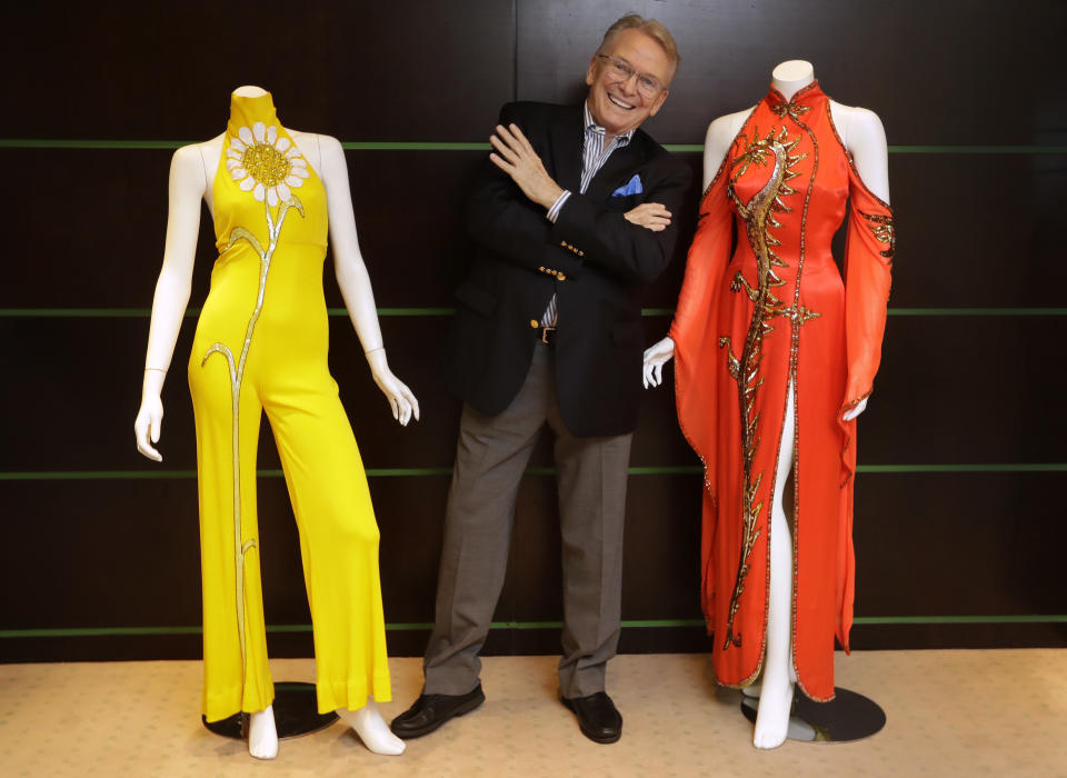 Fashion and Costume Designer Bob Mackie poses with two of his iconic designs, a scarlet red satin gown worn by Cher in 1975, right, and a marigold jersey jumpsuit worn by Cher between 1971-1976, left, in London, Thursday, Aug. 16, 2018. The gowns are estimated at 3,000-5,000 US Dollars ( 2,363-3,938 UK Pounds) each will be auctioned in the 'Property from the Collection of Bob Mackie' sale by Julien's Auctions in Los Angeles on Nov. 17. (AP Photo/Kirsty Wigglesworth)