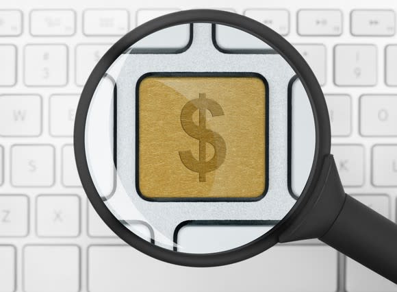 A magnifying glass over a white computer keyboard enlarging a single gold key with a dollar sign on it.