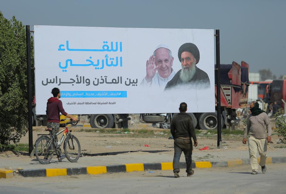 Los iraquíes caminan junto a un cartel que anuncia la próxima visita del Papa Francisco y una reunión con un venerado líder musulmán chiíta, el gran ayatolá Ali al-Sistani