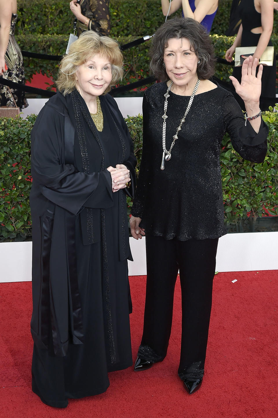 Lily Tomlin and Jane Wagner standing on a red carpet. Lily is wearing a black sequined outfit, and Jane is in a long black coat. Lily is waving