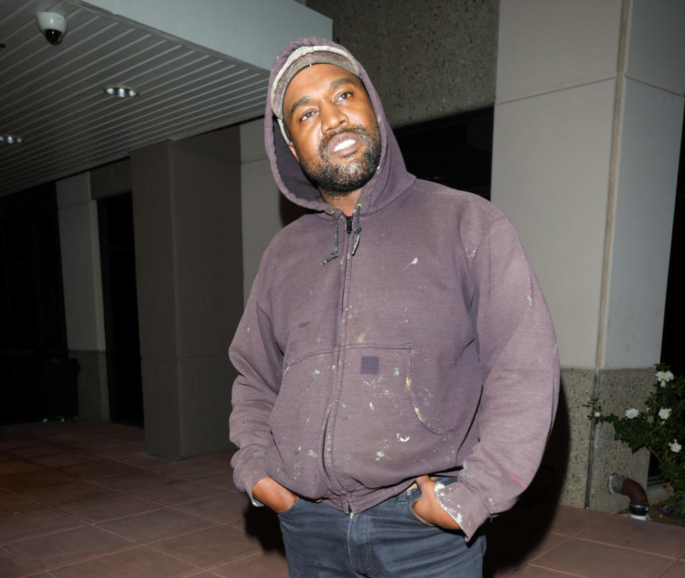 Kanye West in a casual hoodie and jeans, standing with hands in his pockets, looking slightly off to the side