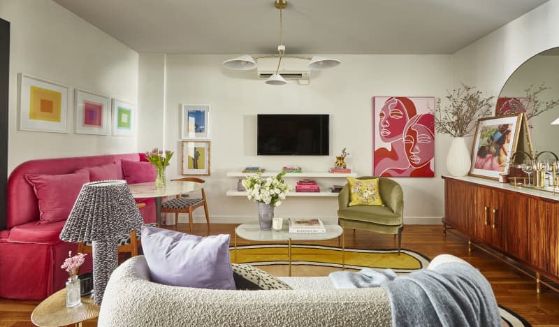 Living room with pink velvet loveseat with small round table, 2 extra chairs for dining. White oval coffee table in front of textured gray loveseat. white shelves under a wall mounted TV with books, a gold vase. Coffee table in foreground with vase of flowers. Green velvet chair with large painting above. Vases of flowers on both tables