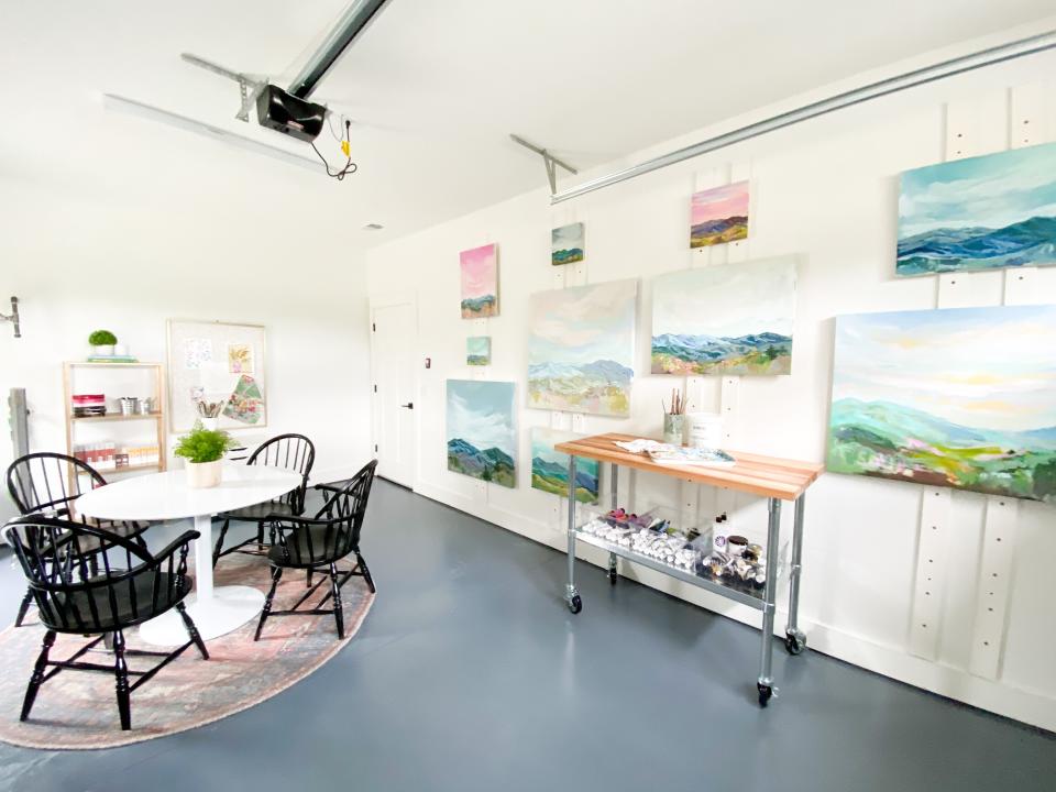 Open, white art studio with scenery paintings hung above work bench and table. 