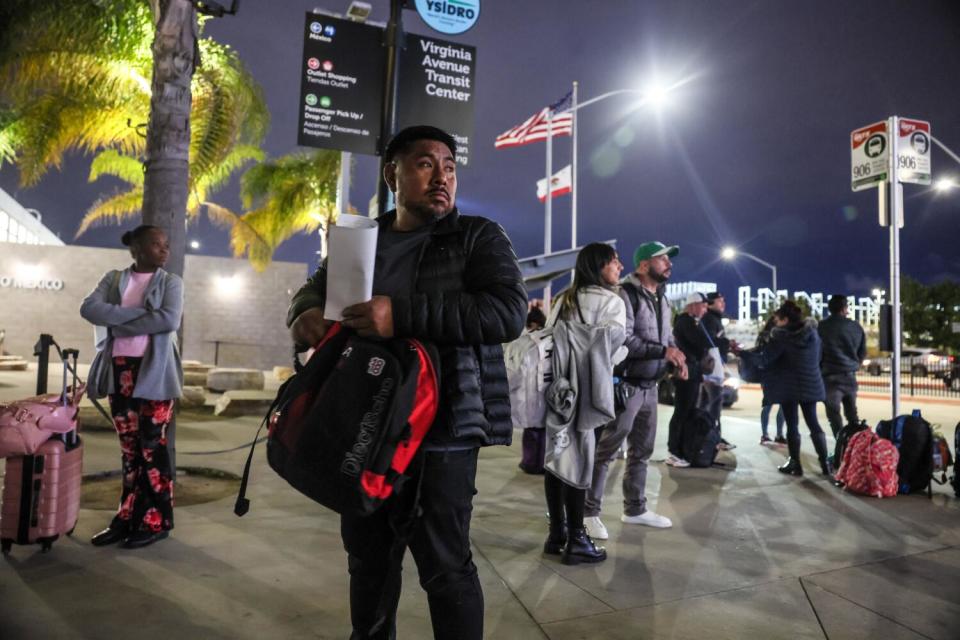 A man in dark clothing holds white papers and a black-and-red backpack near other people with luggage