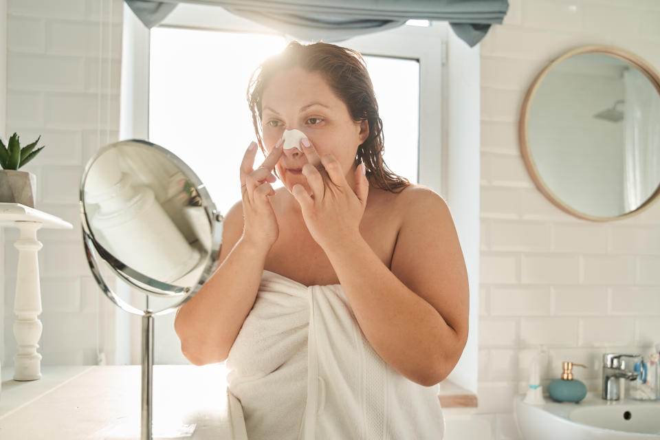 Oversize woman applying nose strips while looking at mirror. Concept of face skin care. Young european girl wearing towel. Interior of bathroom in modern apartment