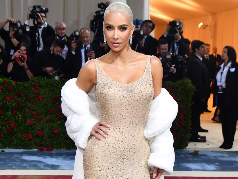 Kim in a nude fitted gown with a scoop neckline and thin straps with chest padding partially seen through the crystal-embellished fabric. She has a white fur stole worn around her arms.
