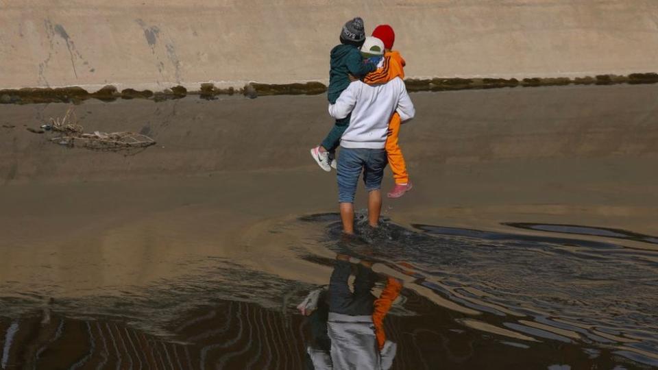 Un hombre cruza un rio en la frontera con dos niños en brazos