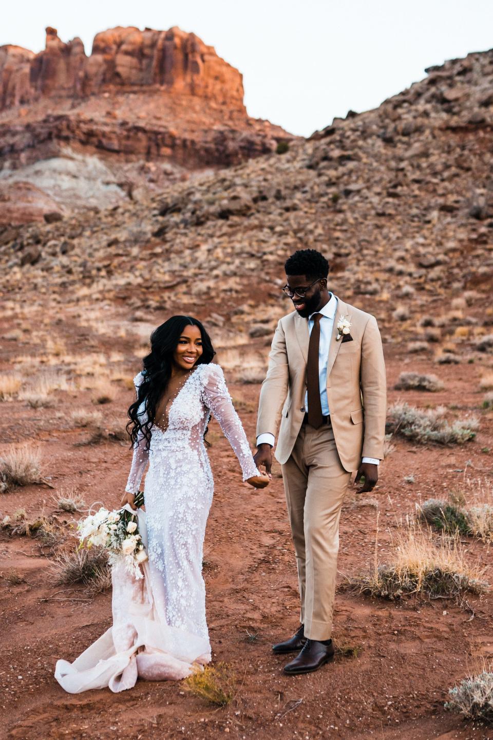A bride and groom smile in their wedding attire in a desert.