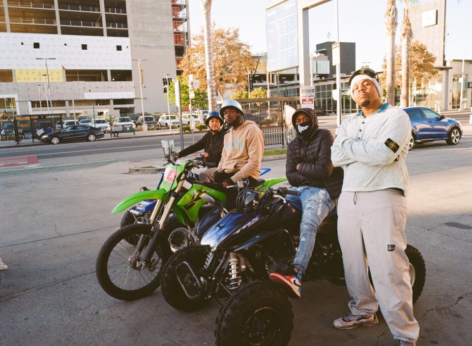 Three men sit on dirt bikes on a city street while a fourth stands next to them, arms folded.