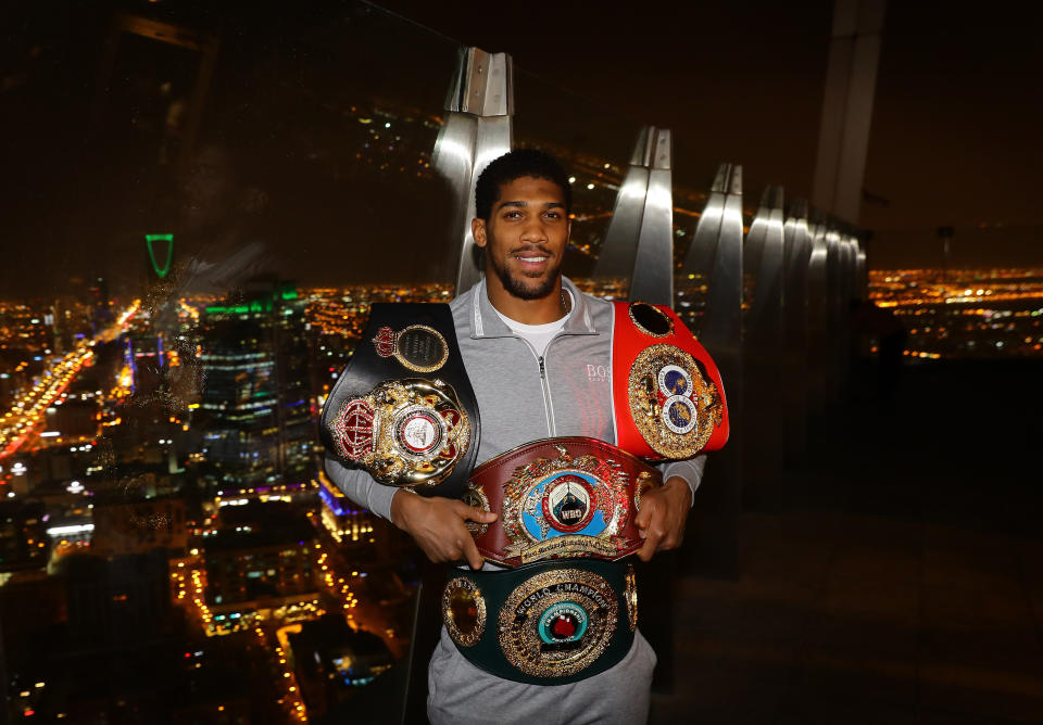 Anthony Joshua with his four title belts in front of a dark city skyline.