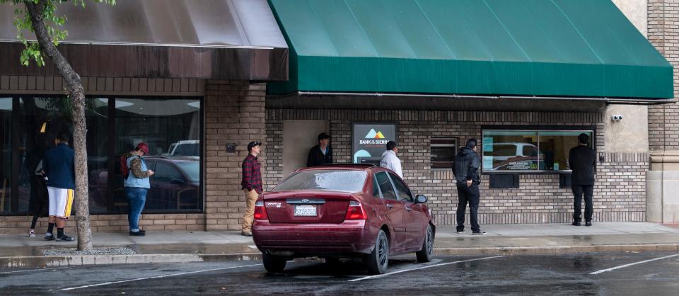 Bank of Sierra customers wait along Main Street for their turn at the walk-up window on Monday, April 6, 2020.