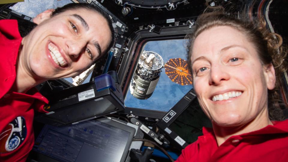 two female astronauts smiling in front of a dome window.  in the back is a spacecraft with solar wings, the Earth in the background