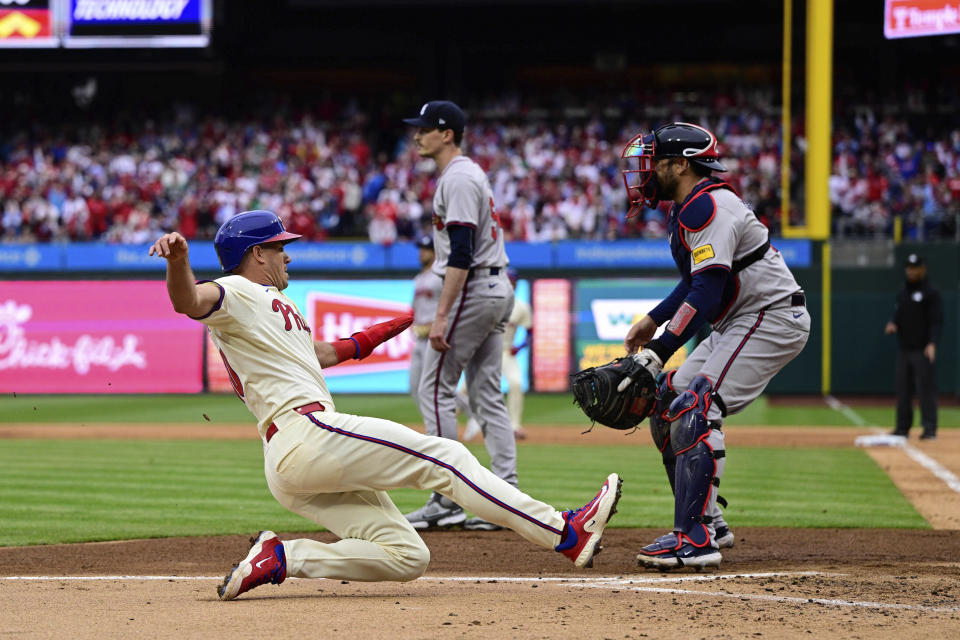 Philadelphia Phillies' J.T. Realmuto, left, slides past Atlanta Braves' Travis d'Arnaud to score a run off a single hit by Bryson Stott during the first inning of a baseball game, Saturday, March 30, 2024, in Philadelphia. (AP Photo/Derik Hamilton)