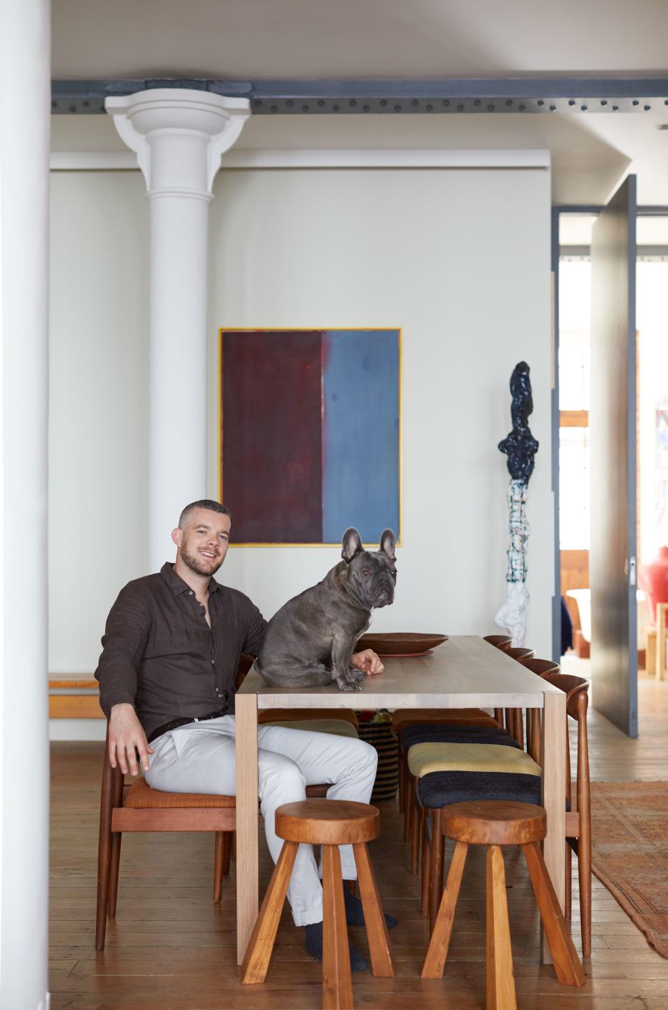 Tovey sits with his sidekick Rocky at the Holborn dining table by e15, which he has mixed with two vintage oak stools from the Peanut Vendor and midcentury chairs that he has collected from a variety of places over the years and had upholstered in a in a selection of Kvadrat fabrics in different colors. In the background is a wooden bench by iconic French designer Charlotte Perriand. Behind him hangs a painting by Matt Connors, next to a sculpture by Rebecca Warren.