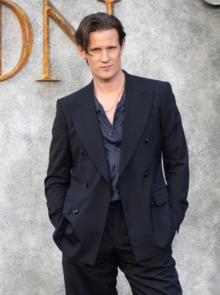 Matt Smith in a dark double-breasted suit with a matching shirt, hands in pockets, at an event. Text on the wall behind him spells "DN"