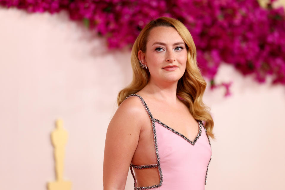 Woman stands on a red carpet wearing a sleeveless pink gown with beaded trim and cut-out sides, with a floral backdrop