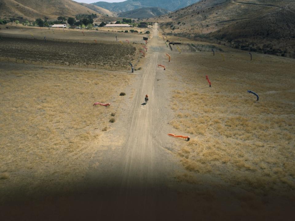 OJ rides along a desert road surrounded by inflatable balloon men while a shadow looms over him.