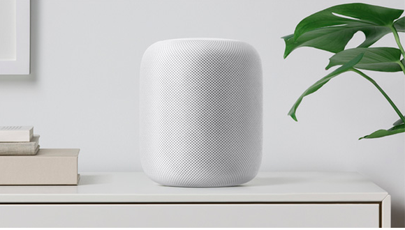 A white Apple HomePod sits on a white table with a white wall behind it.