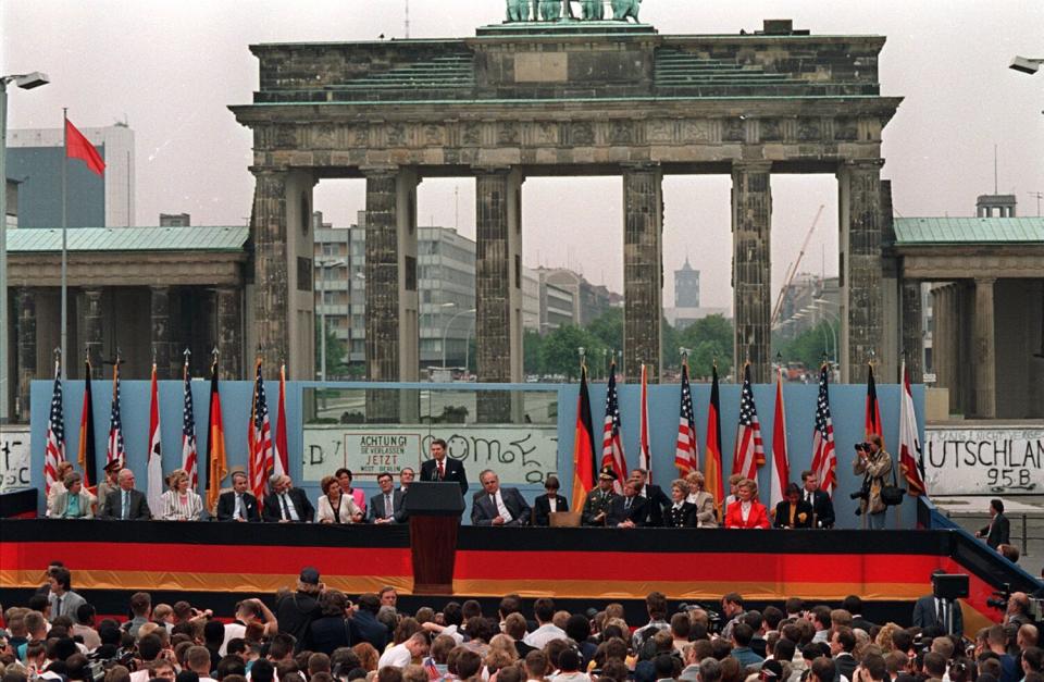 A large monument with six pillars is the backdrop for a man in a suit and tie speaking in front of a row of seated people