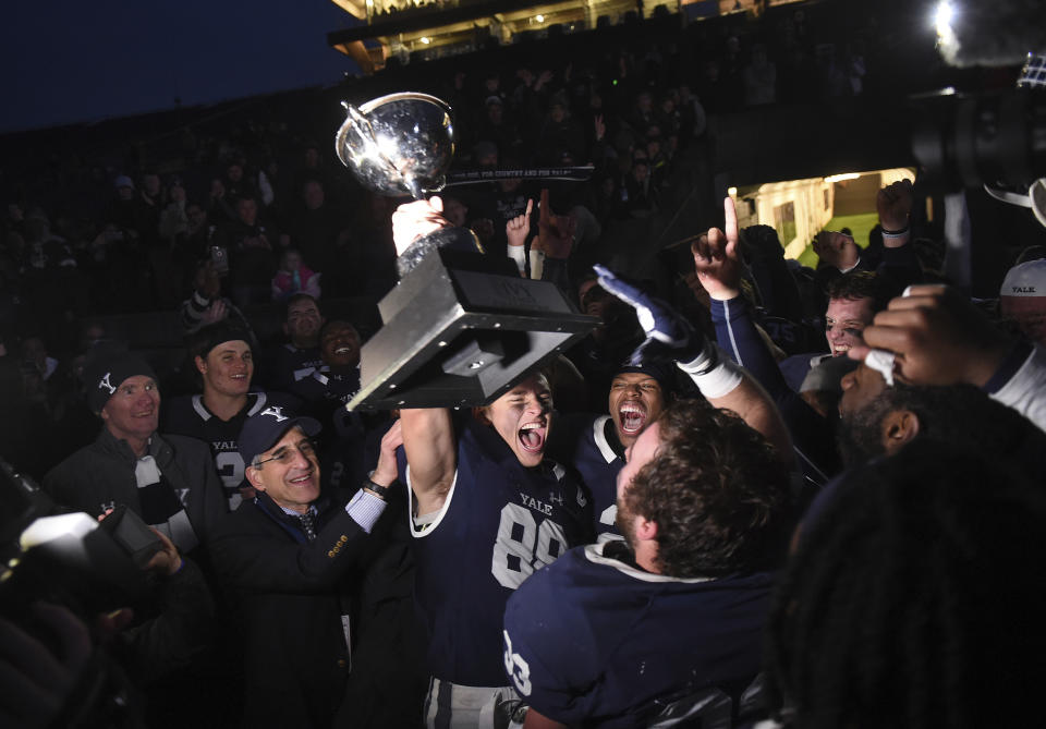 Yale's JP Shohfi, center, holds the Ivy League Championship trophy during a celebration with teammates after their 50-43 double overtime victory against Harvard in an NCAA college football game at the Yale Bowl, Saturday, Nov. 23, 2019, in New Haven, Conn. At left is Yale University president Peter Salovey. (Arnold Gold/New Haven Register via AP)