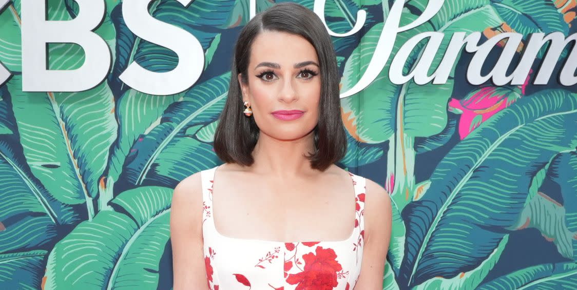 lea michele, a woman stands looking at the camera with a neutral facial expression, she has short brown hair to her shoulders and wears a white dress with red flowers on